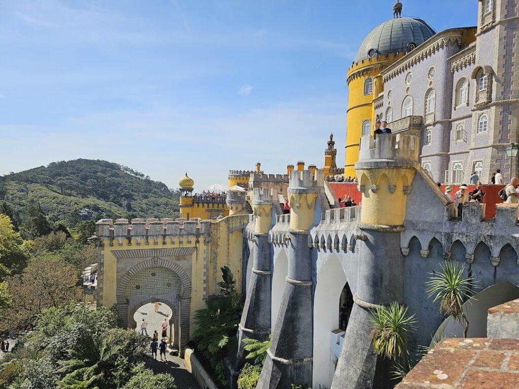 Panoramic view of the vibrant and eclectic architecture of Pena Palace in Sintra, Portugal, with tourists exploring its colorful red, yellow, and tiled façades — a highlight of Yes, You Deserve’s private tuk tuk tours.