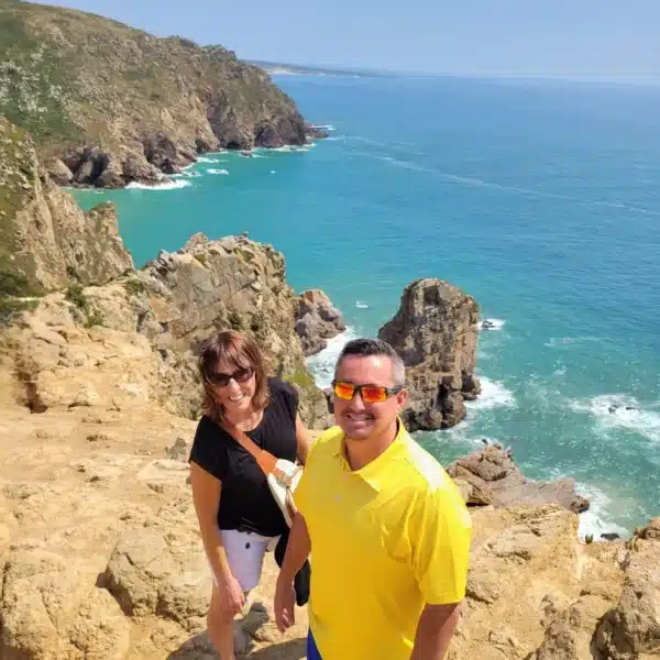 A smiling couple poses at the edge of a dramatic cliff at Cabo da Roca, the westernmost point of continental Europe, with the bright blue Atlantic Ocean and rugged coastline stretching out behind them during a Yes, You Deserve tour.
