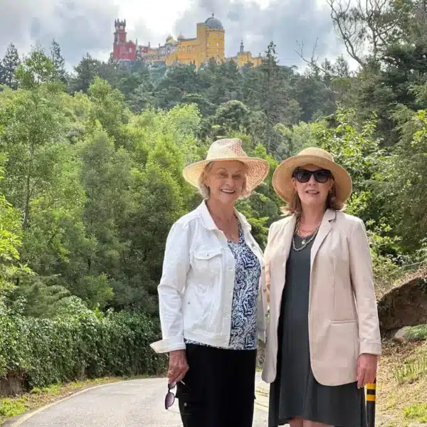 Two women smile during a guided tour with Yes, You Deserve, standing on a scenic road surrounded by forest, with the colorful and majestic Pena Palace visible in the background atop the Sintra hills.