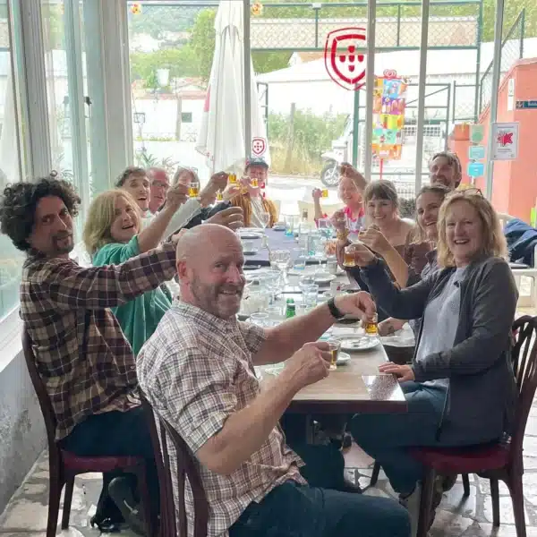 A joyful group of tourists from a Yes, You Deserve tour raises their glasses for a toast during a traditional Portuguese lunch in Sintra. Seated around a long table, they celebrate the experience with smiles and good company inside a cozy local restaurant.