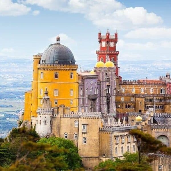 A vibrant view of Pena Palace in Sintra, Portugal, showcasing its colorful architecture with yellow, red, and purple towers perched on a hilltop. A must-see highlight on the Yes, You Deserve tour, offering breathtaking views over the surrounding landscap