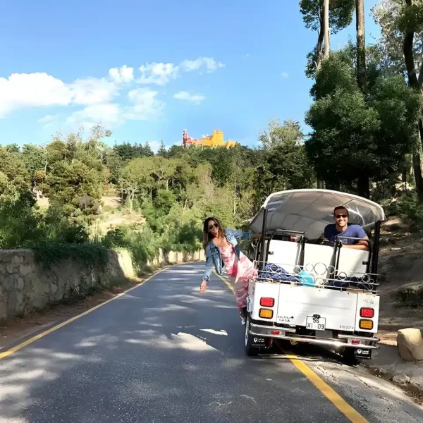 A white tuk-tuk from Yes, You Deserve drives through a scenic forest road in Sintra, with smiling tourists enjoying the ride. In the distance, the colorful Pena Palace peeks through the trees under a bright blue sky.